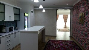 A compact kitchen with white cabinets, a microwave, and a refrigerator. There is a small island in the center, and a colorful rug on the floor. - MANIJA Hostel (Samarkand)