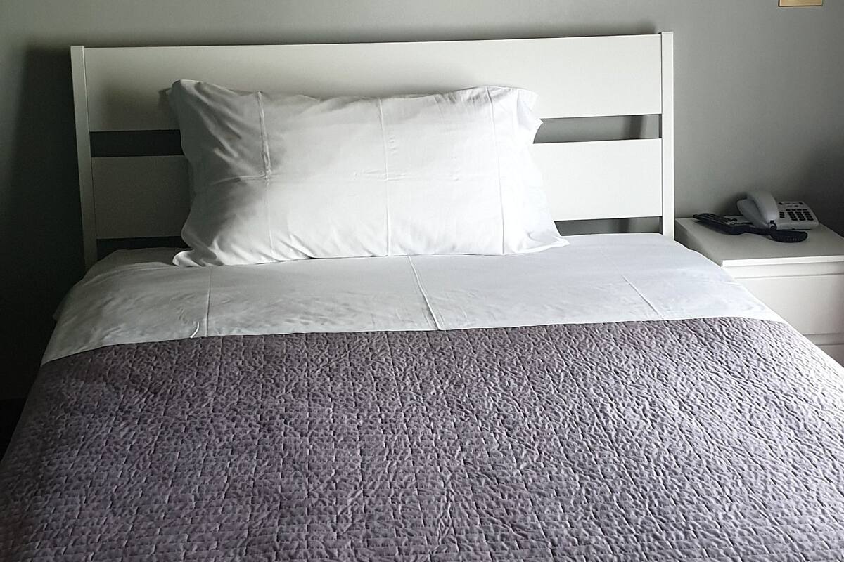 A neatly made bed with a grey quilt, a white headboard, and a wall-mounted artwork of abstract floral designs.
