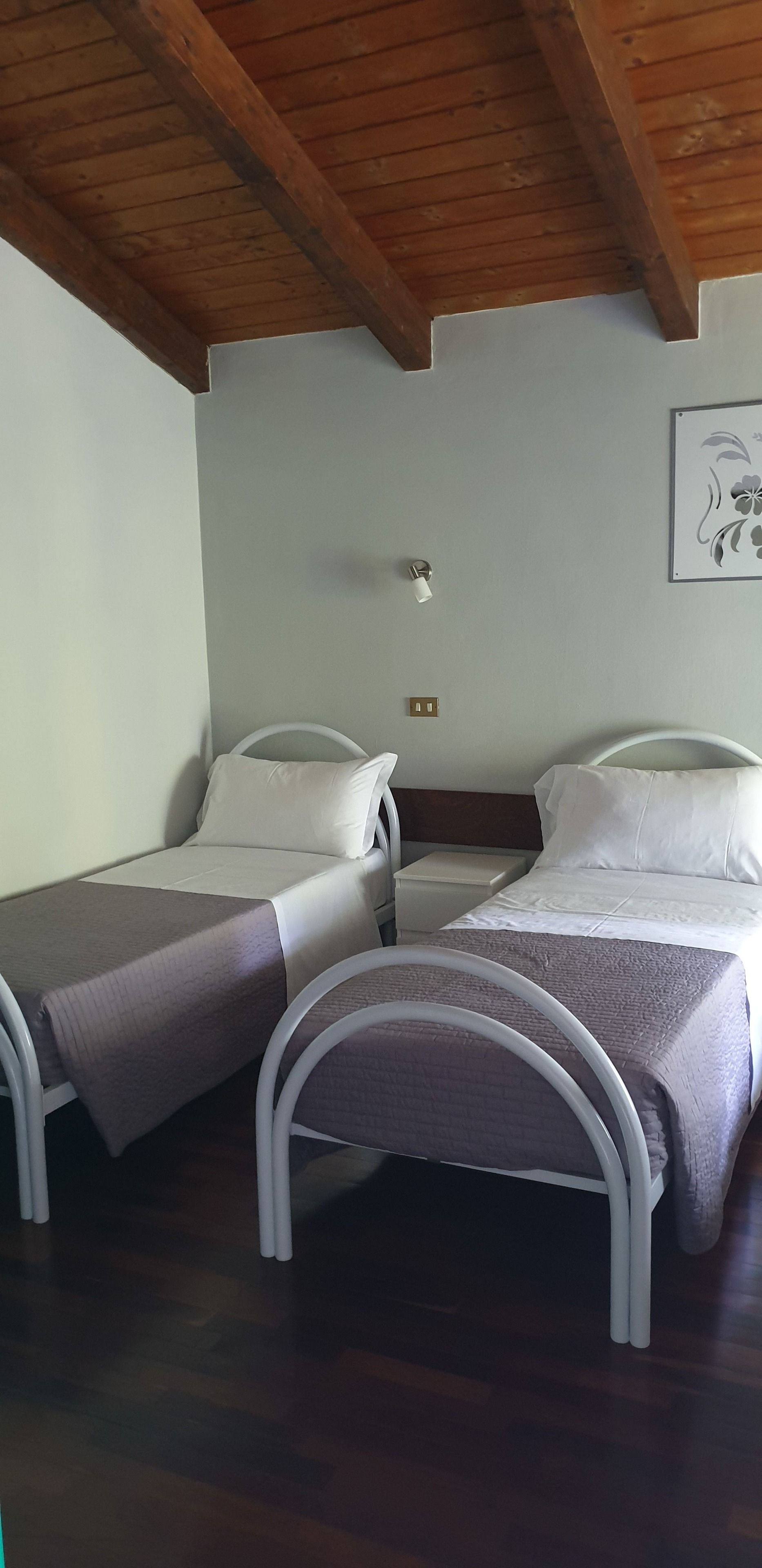 A room with two single beds, wooden ceiling beams, a small bedside table, and a wall-mounted light fixture.