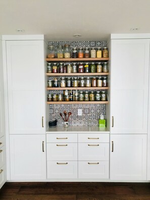 Un garde-manger de cuisine avec des armoires blanches, de nombreuses étagères remplies de bocaux en verre et un revêtement mural décoratif.