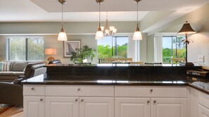 A kitchen with a black countertop, white cabinets, and pendant lights. There is a dining area with a table and chairs, and a view of greenery outside.