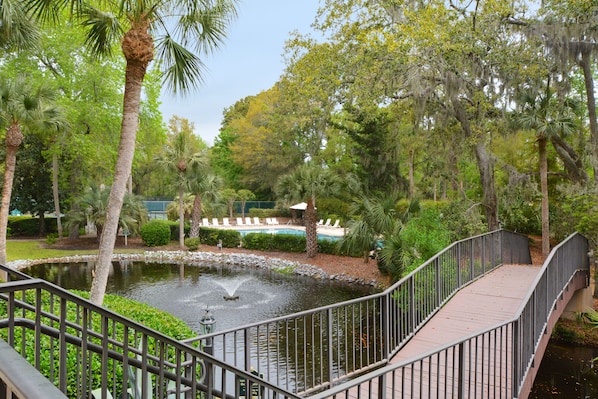A bridge over a pond with a fountain, surrounded by lush greenery and a pool area in the background.