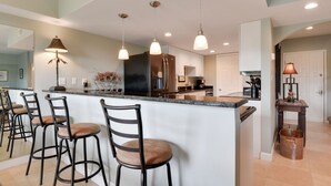 A modern kitchen with a central island, bar stools, pendant lighting, and a large refrigerator.