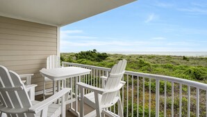 A covered outdoor seating area with white chairs and a table, overlooking a beach with green vegetation. - Oceanfront Condo with Ocean Views (Wrightsville Beach)