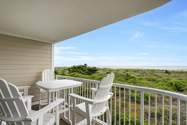 A covered outdoor seating area with white chairs and a table, overlooking a beach with green vegetation.