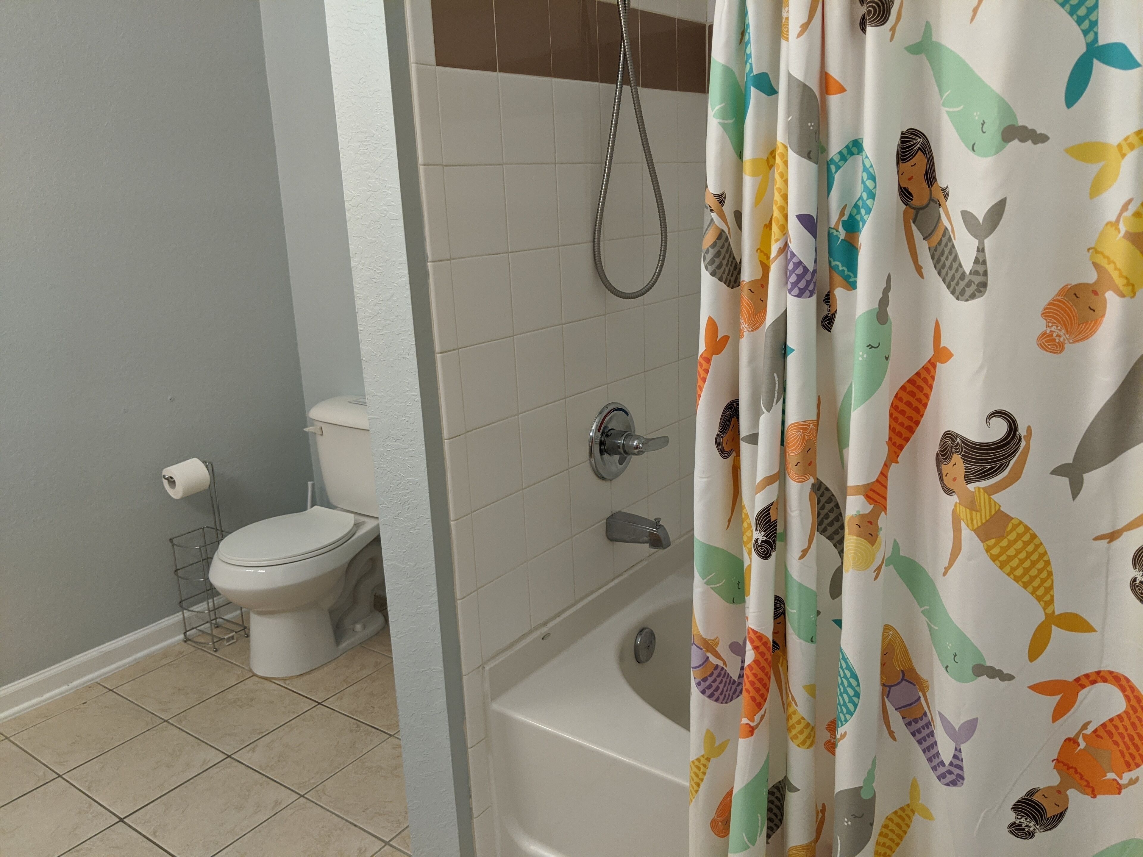 A bathroom with a toilet, a shower with a curtain featuring mermaid designs, and tiled walls.