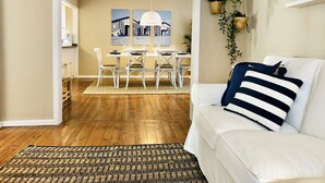 Ein Wohnzimmer mit weißem Sofa, einem gemusterten Teppich, einem Essbereich mit Tisch und Stühlen sowie einer hängenden Pflanze.