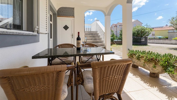 Un patio techado al aire libre con una mesa y sillas de mimbre, una botella de vino y vistas a una escalera que lleva a un edificio.