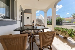 A covered outdoor patio with a table and wicker chairs, a bottle of wine, and a view of a staircase leading to a building.
