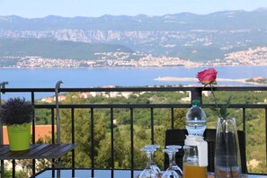 A balcony with a table set for two, a rose, and a scenic view of a coastal town. - Apartment Baresic Roberta (Silo - island Krk)
