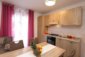 A modern kitchen with wooden cabinets, a dining table with fruit, and a sofa in the background. - Apartment Mirjana rozi (Silo - island Krk)