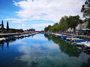 Une rivière paisible, bordée de chaque côté par des bateaux à quai, et entourée d’arbres et de bâtiments.