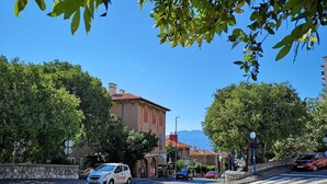A street view with a white car parked on the side, a building with a red roof, and trees in the background. - Krimeja apartment close to Pecine beach and center (Rijeka)