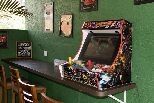 A bar area with a retro arcade machine, framed motorcycle and car posters, and wooden chairs. - Pousada Du Valle (Embu das Artes)
