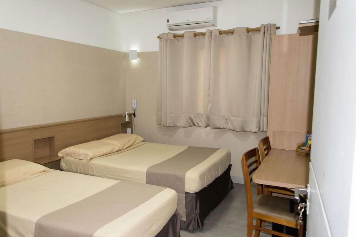 A hotel room with two beds, a wooden table, a chair, an air conditioning unit, and a curtain.