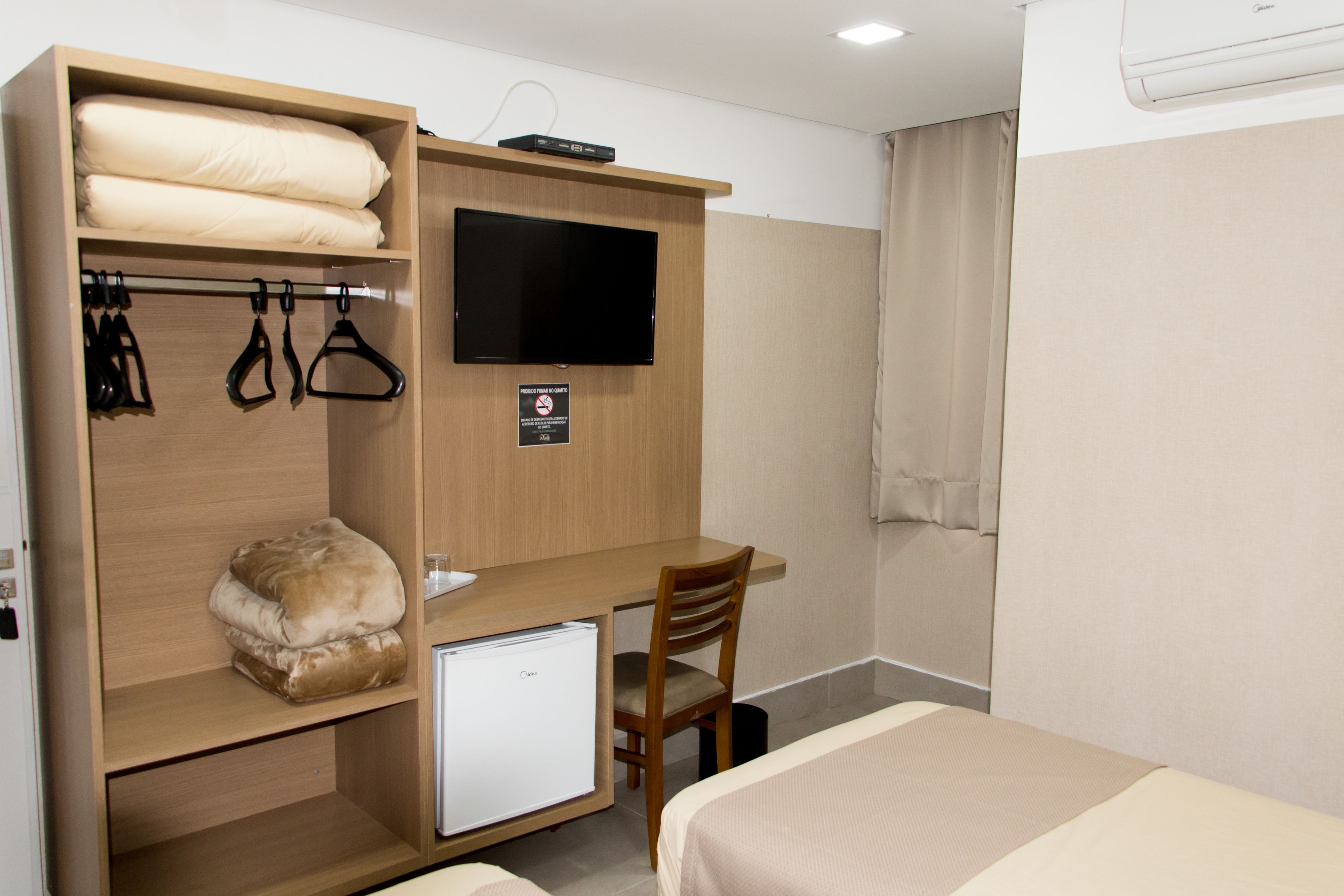A compact hotel room with a bed, a desk with a chair, a television, a mini-fridge, and a wardrobe with hanging items.