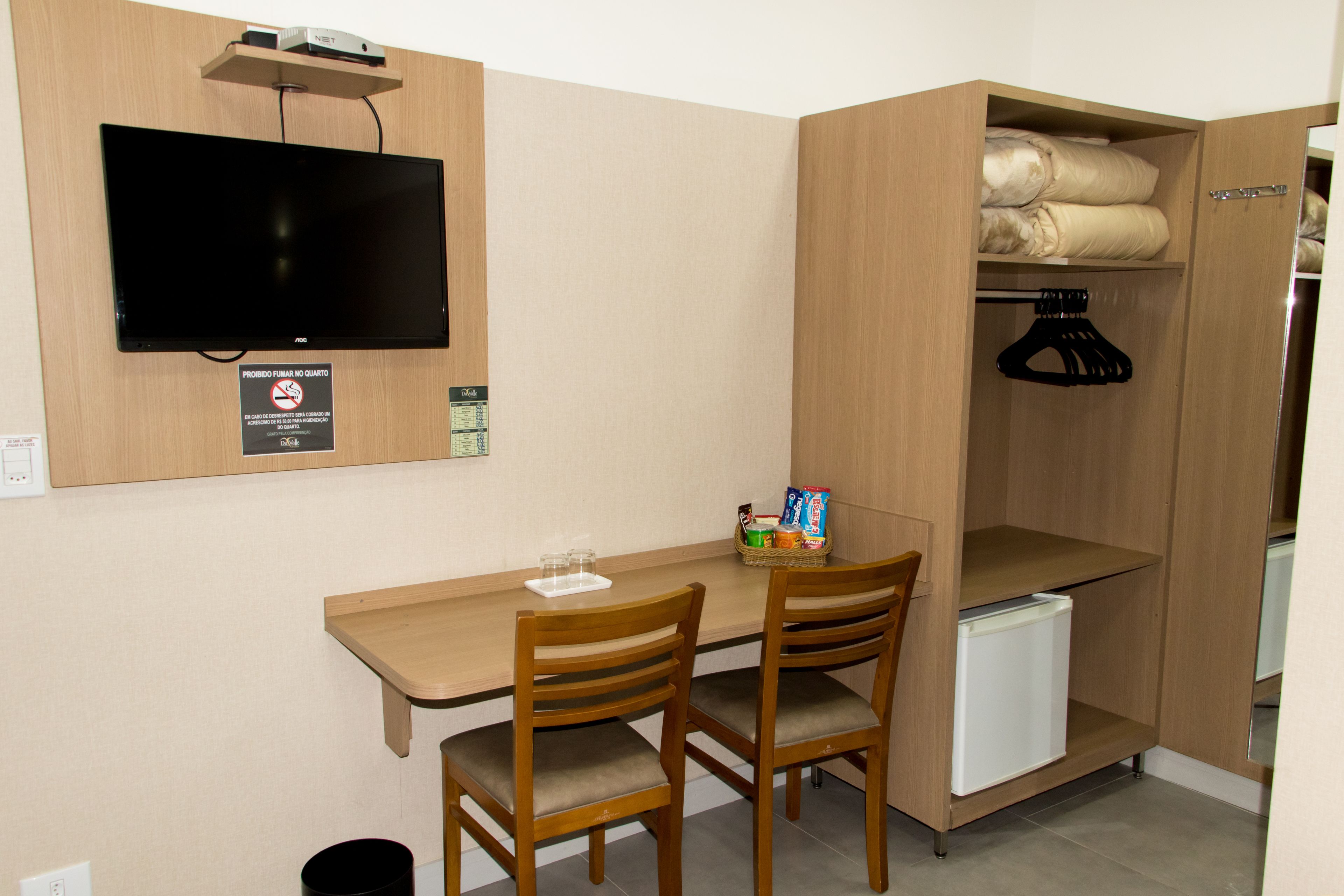 A compact room with a wooden desk, two chairs, a television mounted on the wall, a small refrigerator, and a wardrobe with hanging space.