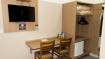A compact room with a wooden desk, two chairs, a television mounted on the wall, a small refrigerator, and a wardrobe with hanging space.