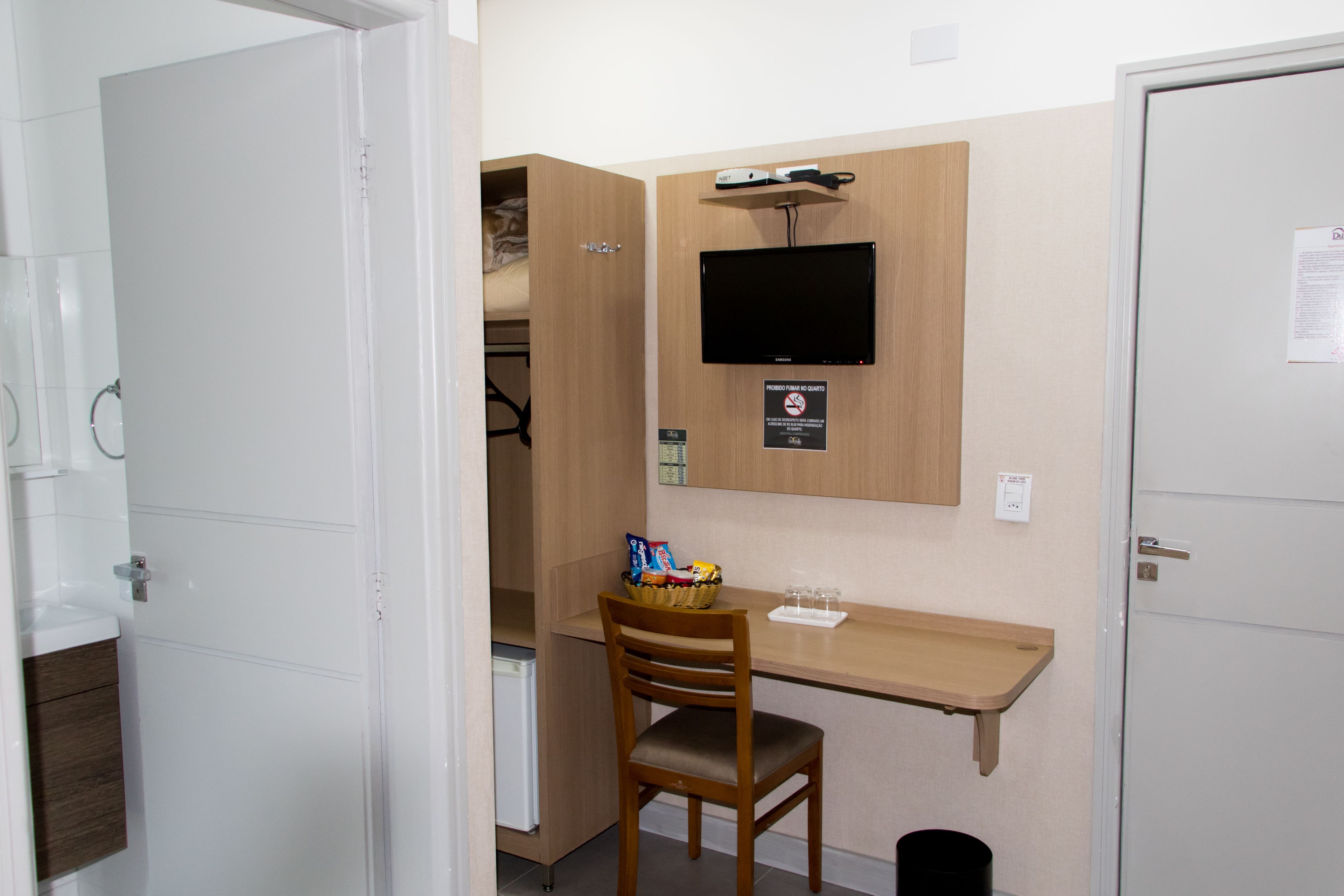 A compact room with a wooden desk, a television mounted on the wall, a small refrigerator, and a chair.