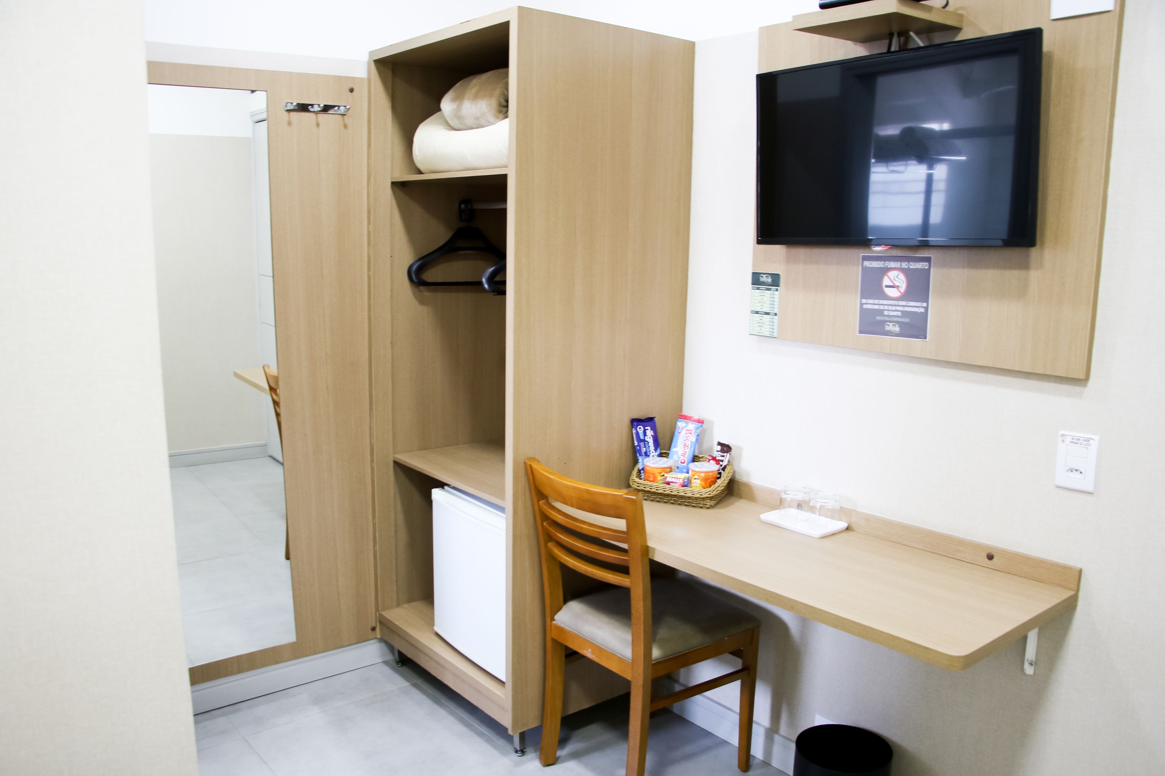 A compact room with a wooden desk, a chair, a television mounted on the wall, and a small refrigerator.