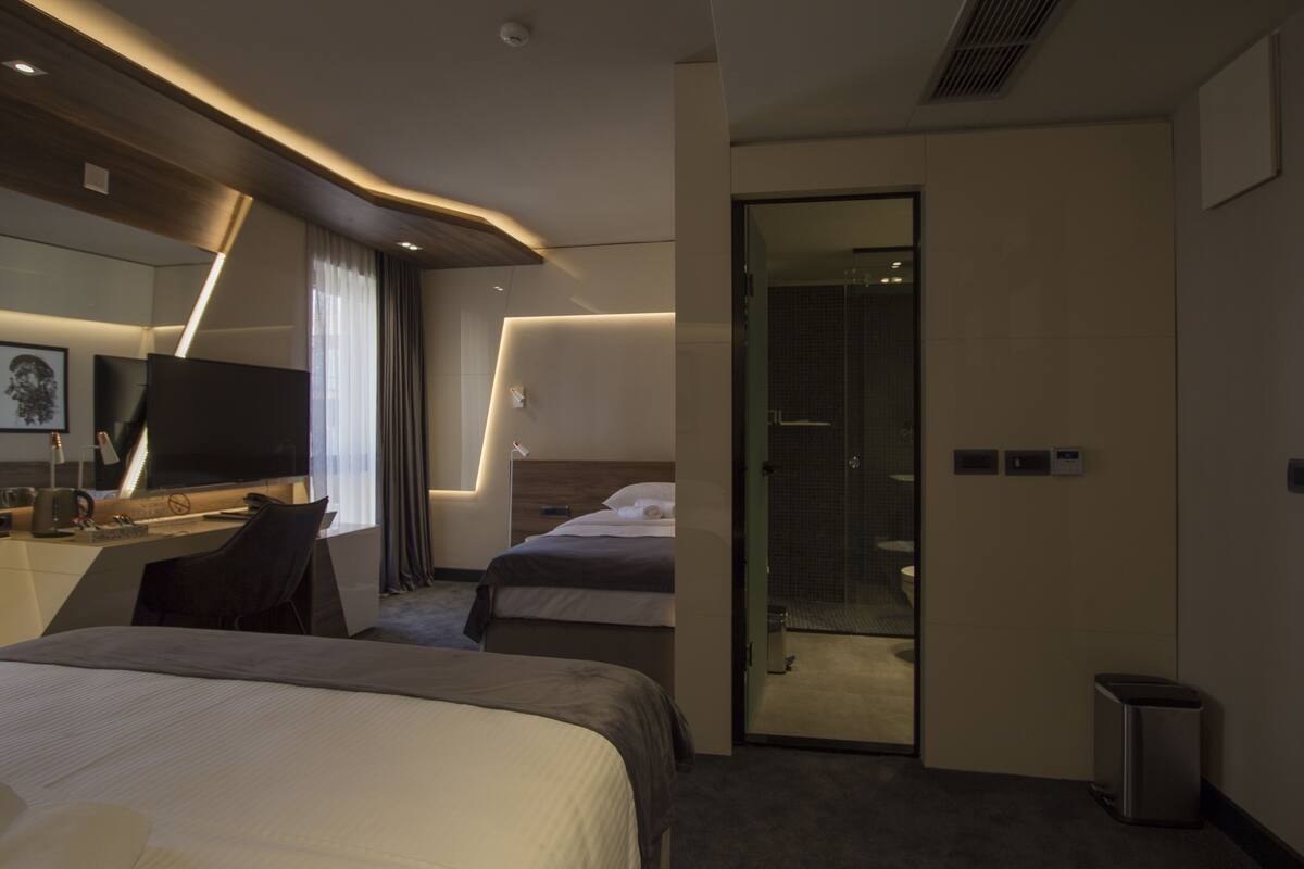 A modern hotel room with a large bed, a desk with a computer, a TV, and a shower area.