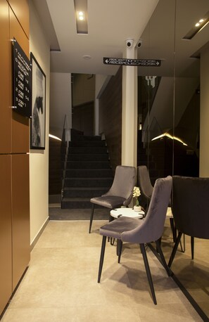 A modern hotel lobby with a reception desk, two grey armchairs, a vase with flowers, and a staircase in the background.