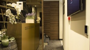 A reception desk with a large potted plant, a flat-screen TV mounted on the wall, and a wooden panel wall in the background. - Garni Hotel Boutique 25h (Niš)