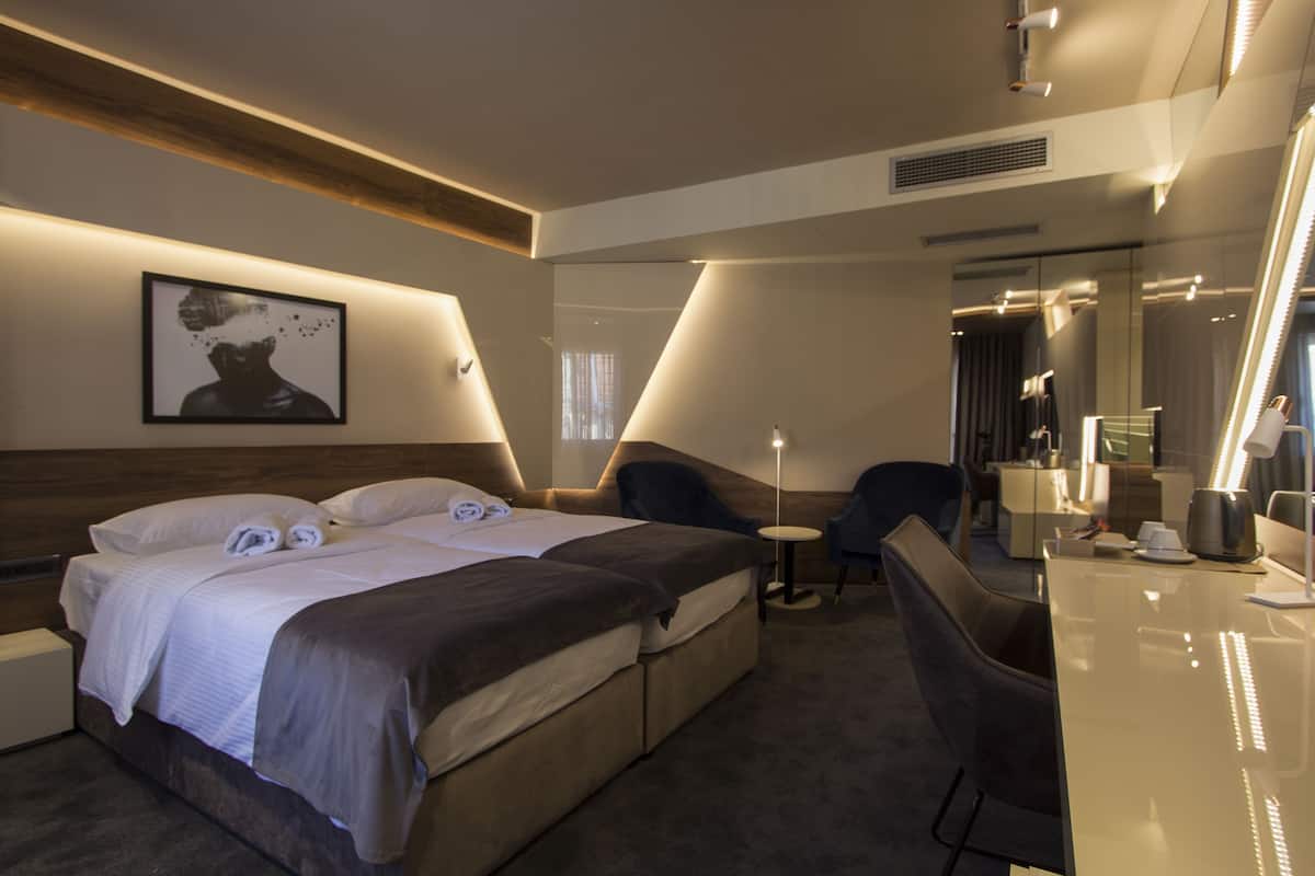 A modern hotel room with a large bed, a desk, and a chair. There is a painting on the wall and a mirror above the desk.