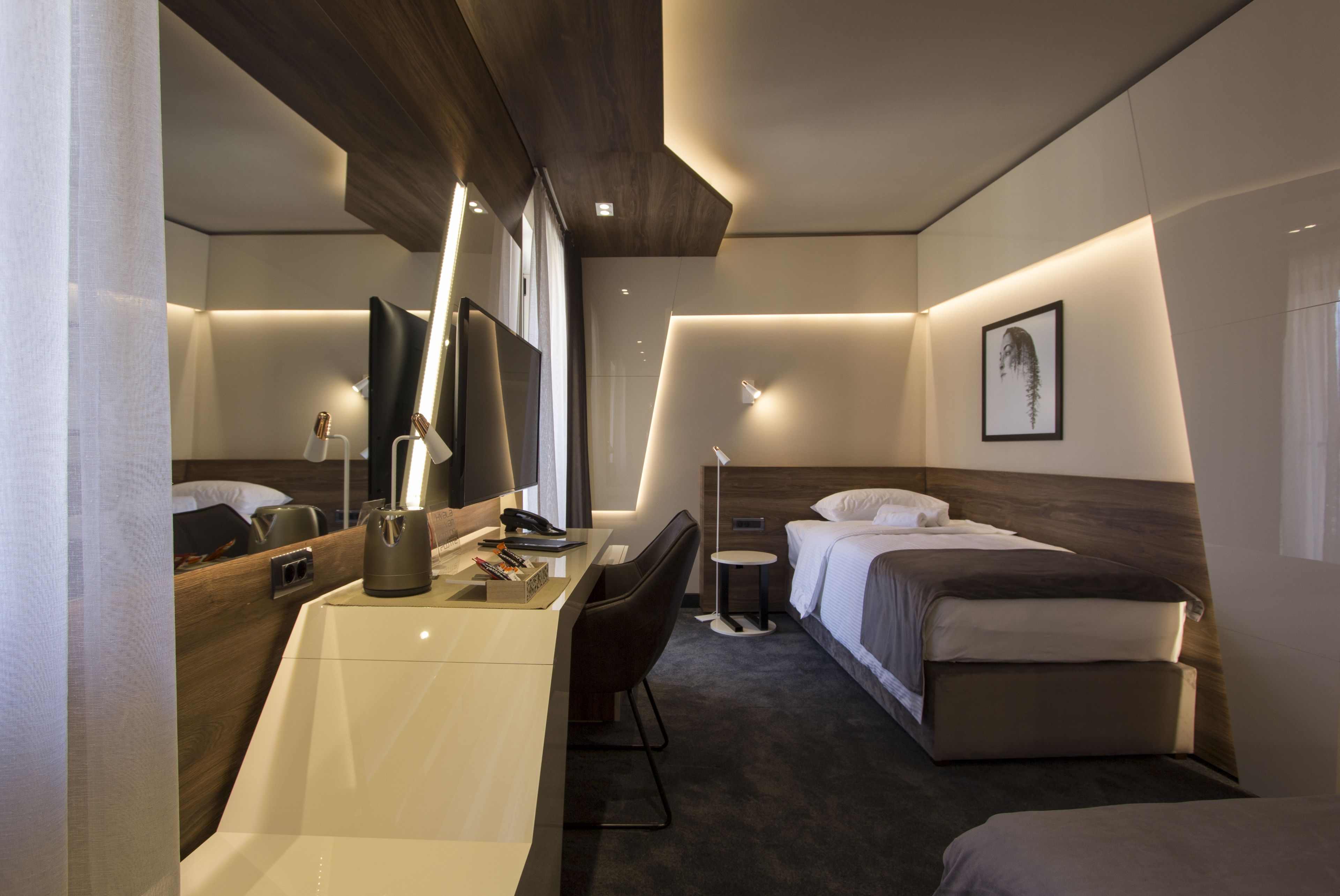 A modern hotel room with a desk, chair, bed, bedside table, lamp, and a large mirror.