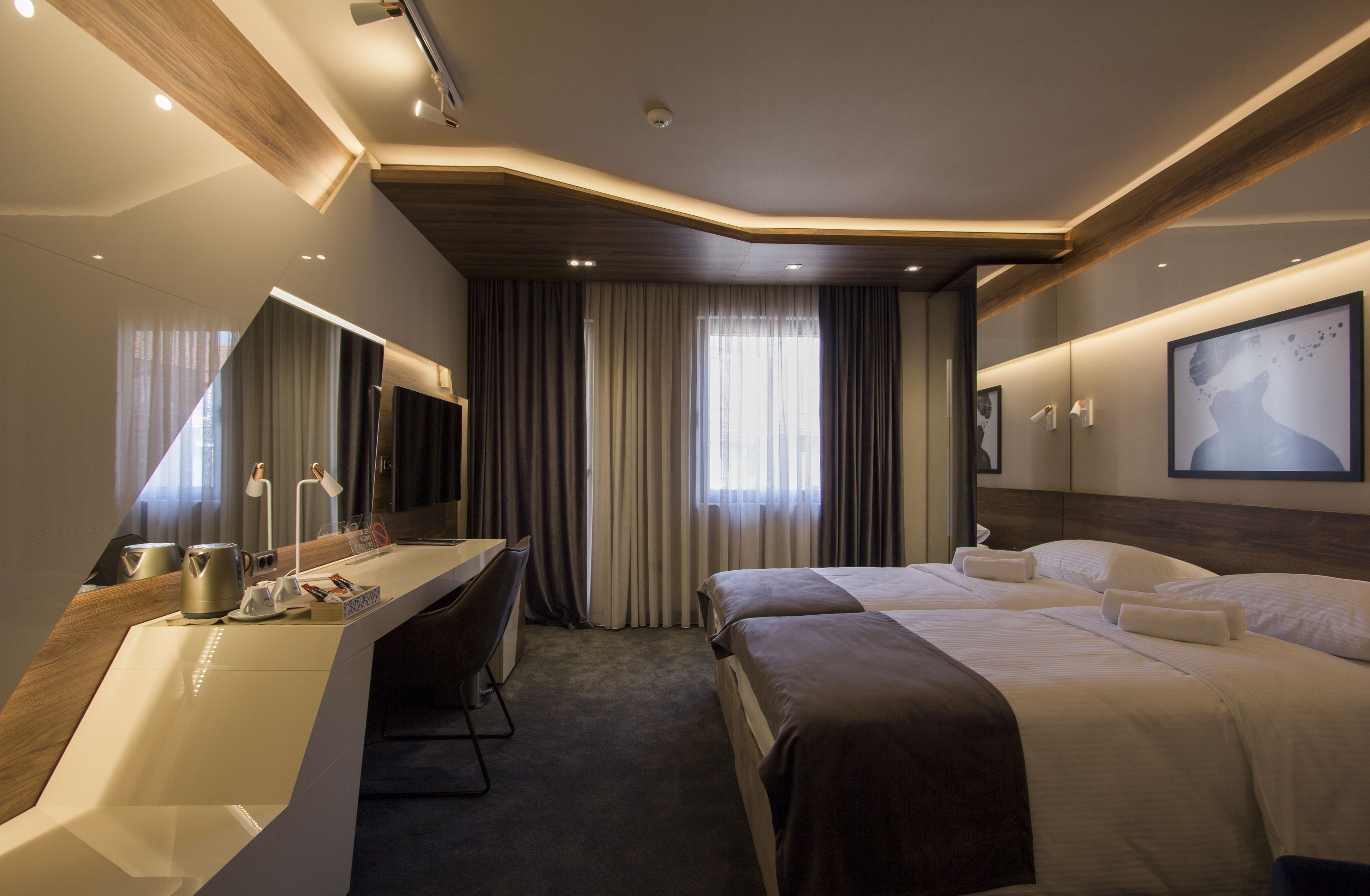 A modern hotel room with a desk, two beds, a large mirror, and a painting on the wall.