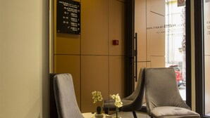 A modern waiting area with two grey upholstered chairs, a small table with a vase of flowers, and a sign listing room numbers. - Garni Hotel Boutique 25h (Niš)