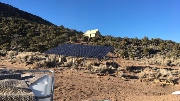 A solar panel array in a dry, hilly landscape with a house in the background. - Hike By Light, Campers Delight. (Fort Garland)