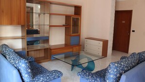 A living room with a glass coffee table, blue patterned sofas, wooden cabinets, and a door. - Bethel Bed and Breakfast (Gela)