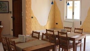 A dining area with wooden tables and chairs, a window with blinds, and a door. - Bethel Bed and Breakfast (Gela)