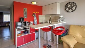 Eine Küche mit roten Schränken, einer weißen Arbeitsplatte, einer Mikrowelle, einer Kaffeemaschine und einer Weinflasche. Zwei rote Barhocker und ein beiges Sofa befinden sich im Raum.