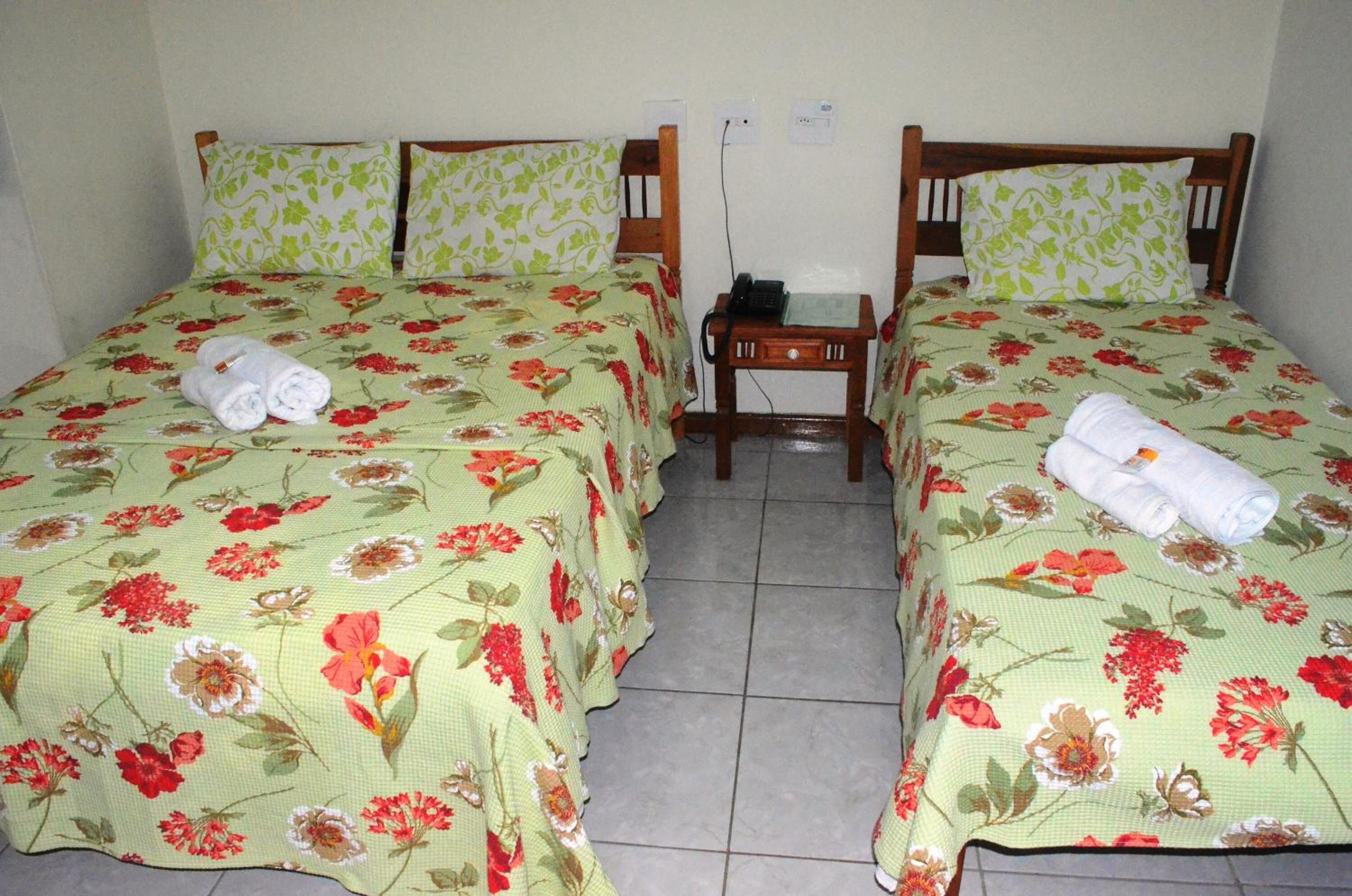 A room with two beds, each with floral bedspreads and matching pillows. There is a small wooden nightstand with a telephone and a book between the beds.