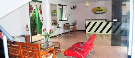 A reception area with a wooden bench, a red chair, a glass table with a vase, and a sign reading "Pousada Brisa do Vale".