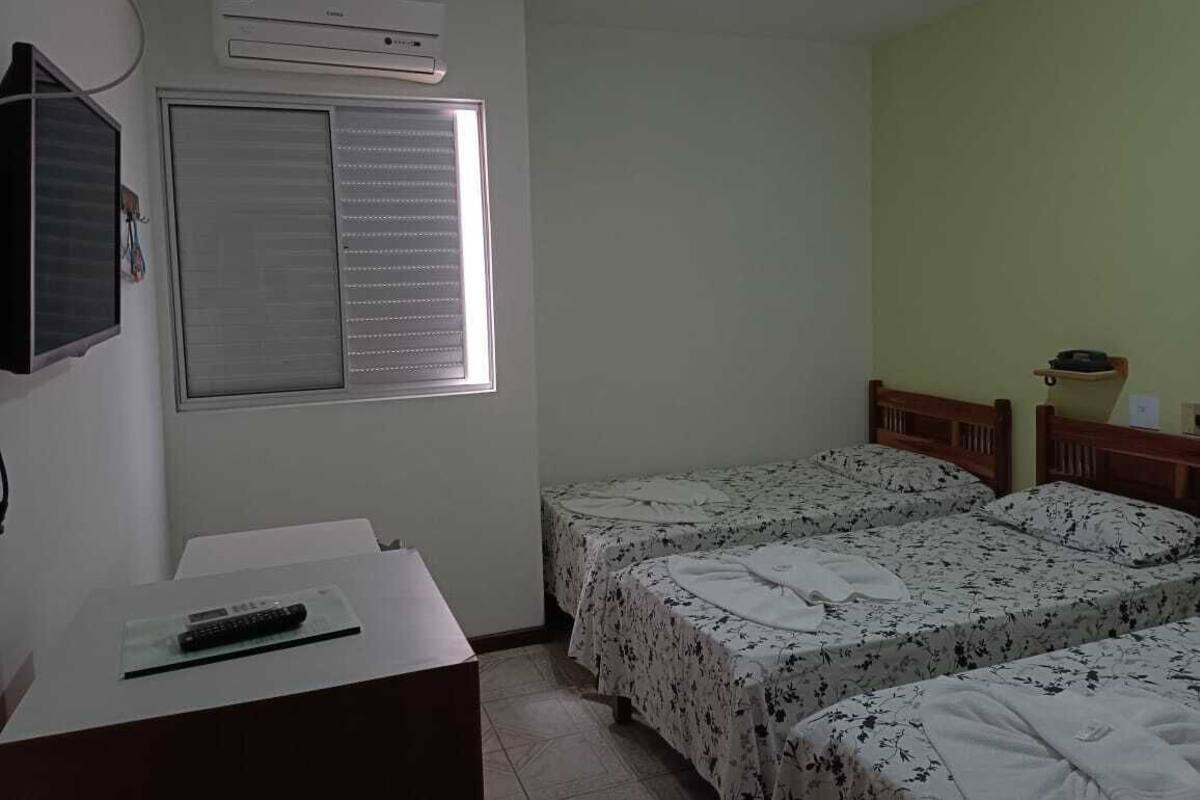 Quarto com duas camas, uma escrivaninha com controle remoto, uma janela com persianas e um aparelho de ar condicionado.