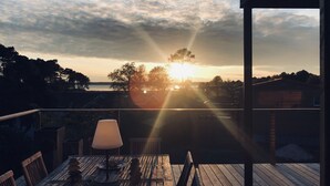 Una terraza de madera con una mesa y sillas, con vista a un atardecer sobre una masa de agua, árboles y una costa lejana.