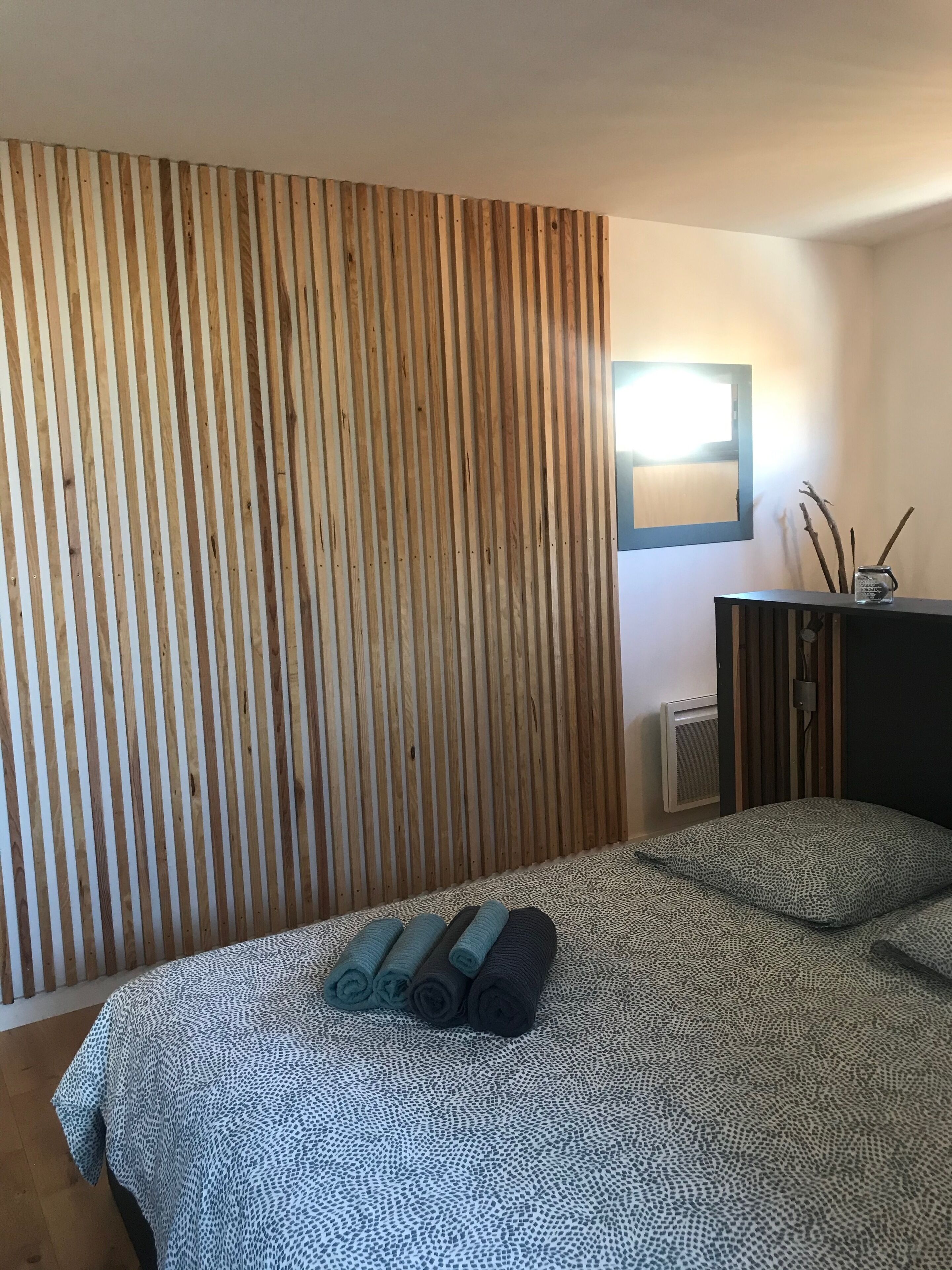 Une chambre avec un mur recouvert de lambris en bois, un lit avec une literie à motifs et une petite table sur laquelle se trouvent une lampe et des branches décoratives.