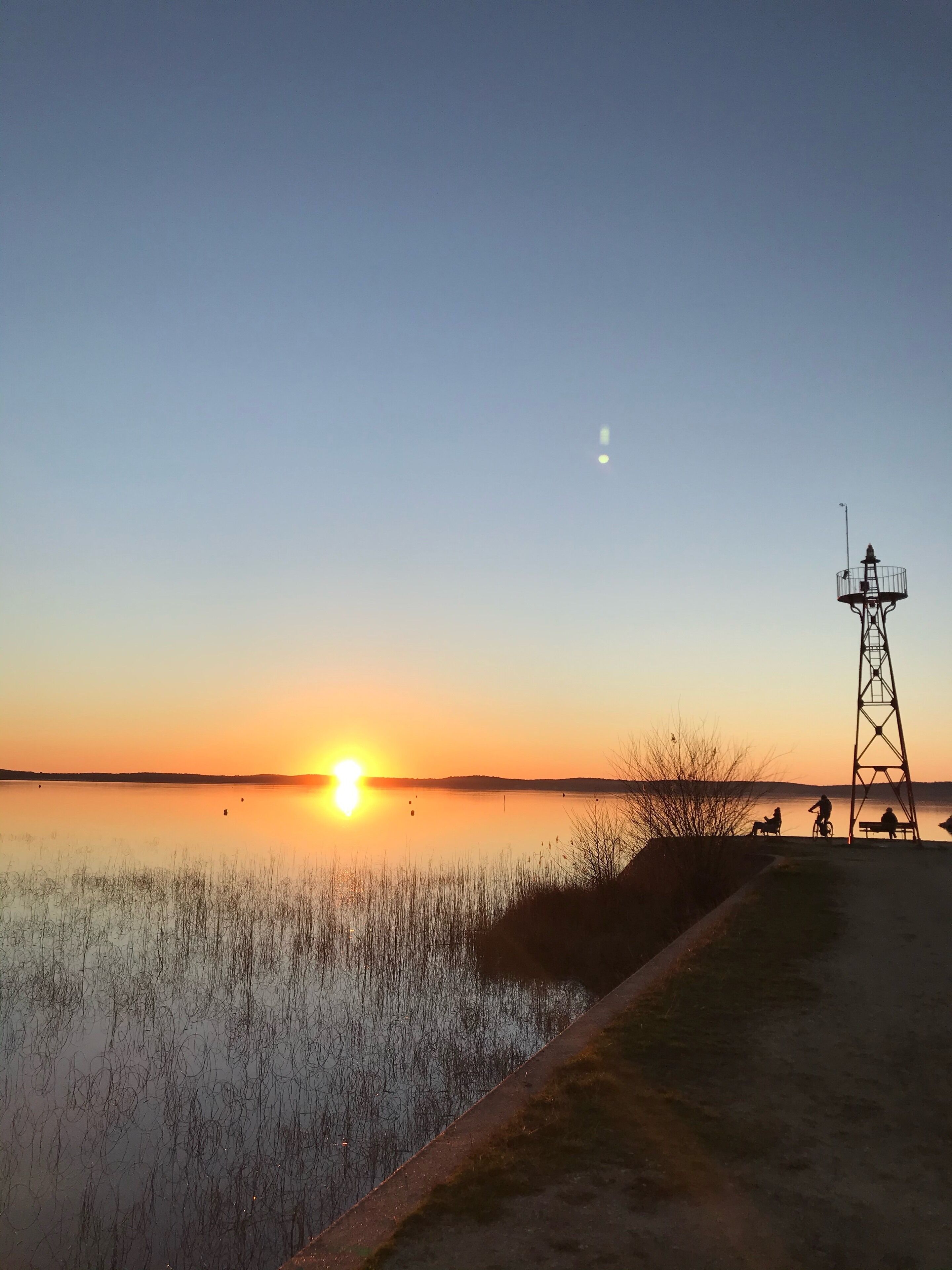 A sunset over a calm body of water with a utility tower and a path along the shore.