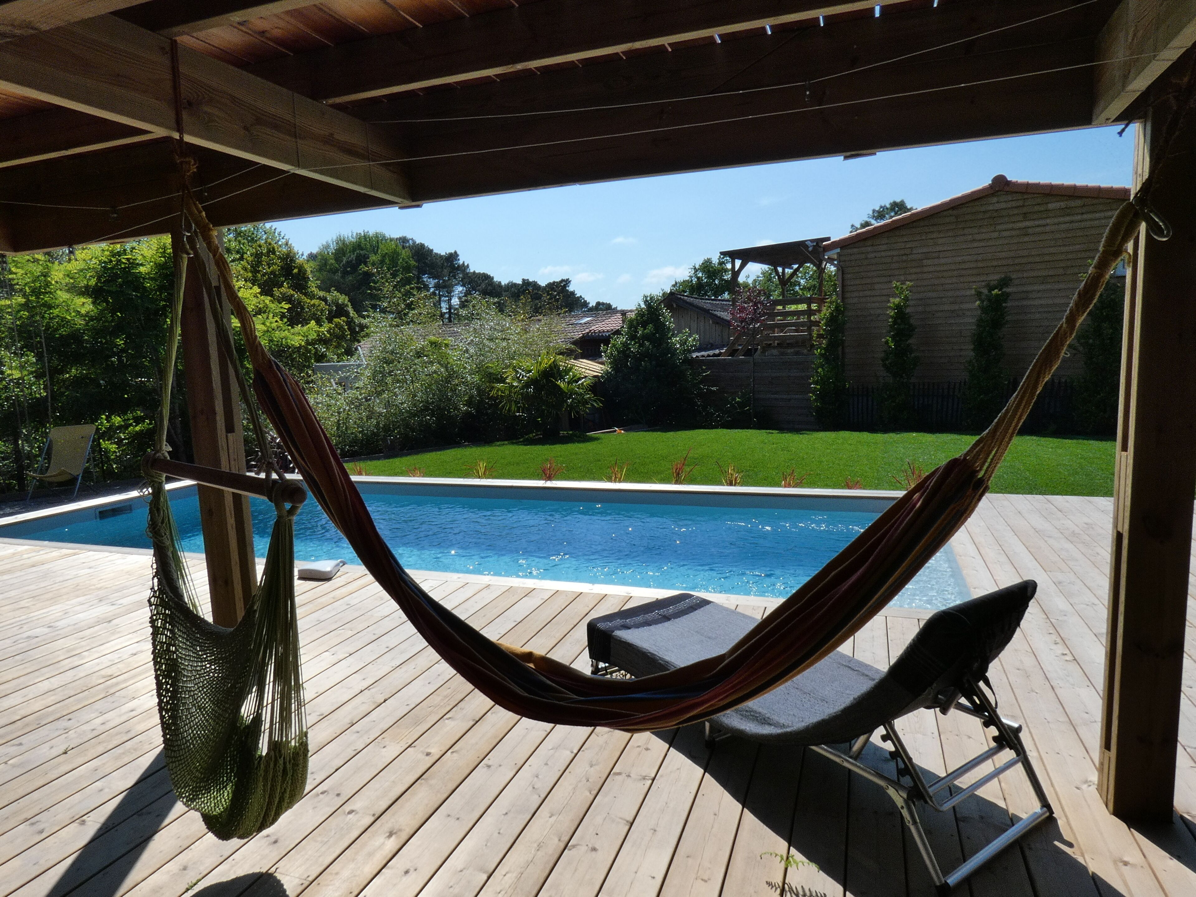 En hængekøje under en træpergola med udsigt over en swimmingpool og en græsplæde.