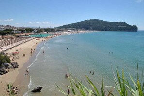 Platja amb ombrel·les i persones, un cel clar i un turó a la distància.