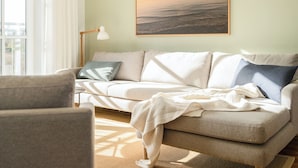 Ein Wohnzimmer mit einem beigen Sofa, einer weißen Lampe und einem Bilderrahmen mit einem Sonnenuntergang über dem Meer.