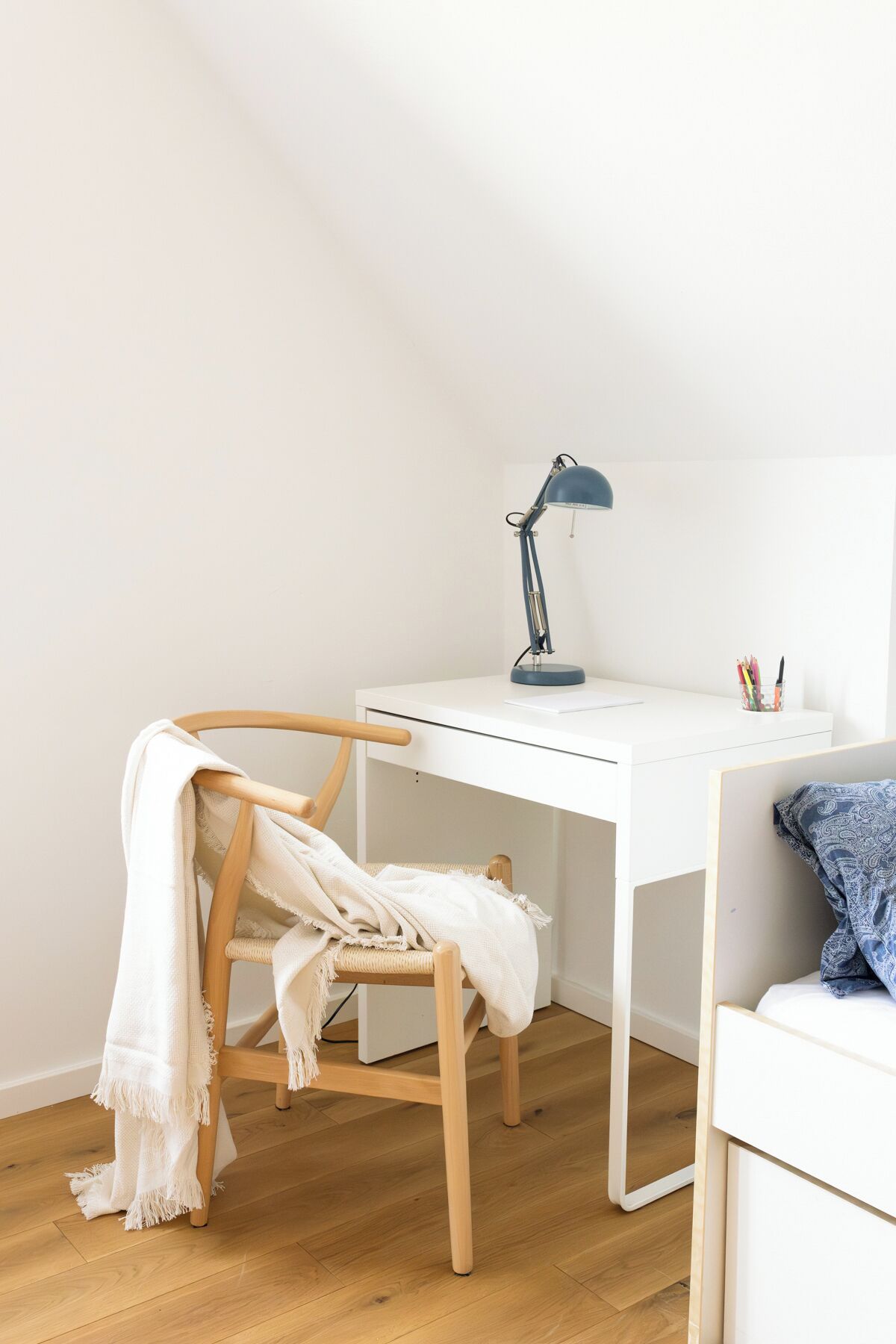Un escritorio blanco con una lámpara de escritorio azul, una silla de madera con una manta blanca y una cama con una colcha azul estampada.