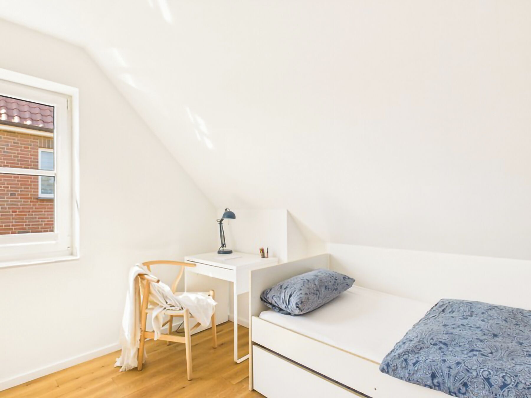 Um quarto estilo loft pequeno e branco, com uma escrivaninha e cadeira, uma cama com manta estampada, e janela com vista para um edifício de tijolos.
