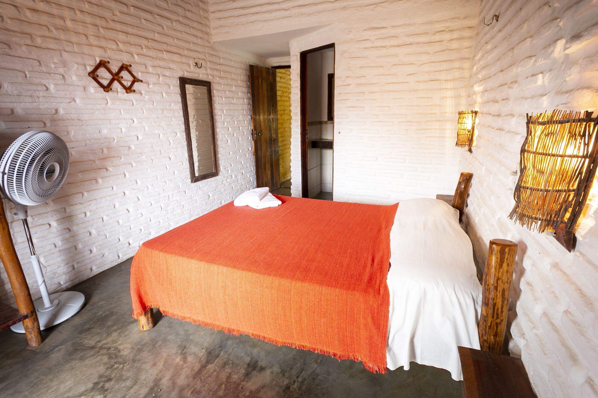 Quarto com cama, ventilador e espelho em parede de tijolos.