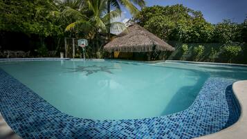 Piscina com borda em mosaico azul, rodeada por vegetação exuberante e uma estrutura de telhado de palha.