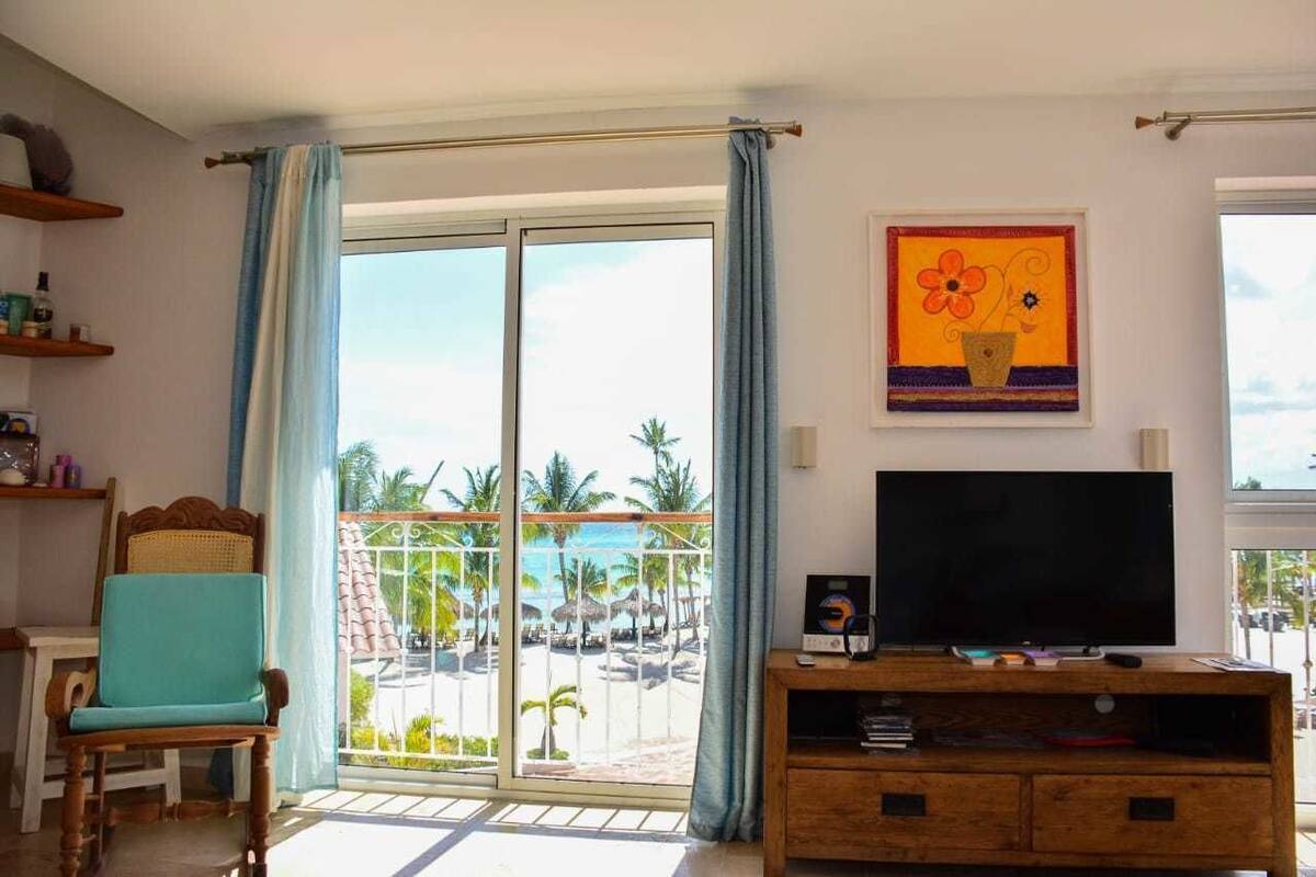 A room with a TV on a wooden stand, a chair, a balcony with a view of palm trees and the sea, and a painting on the wall.