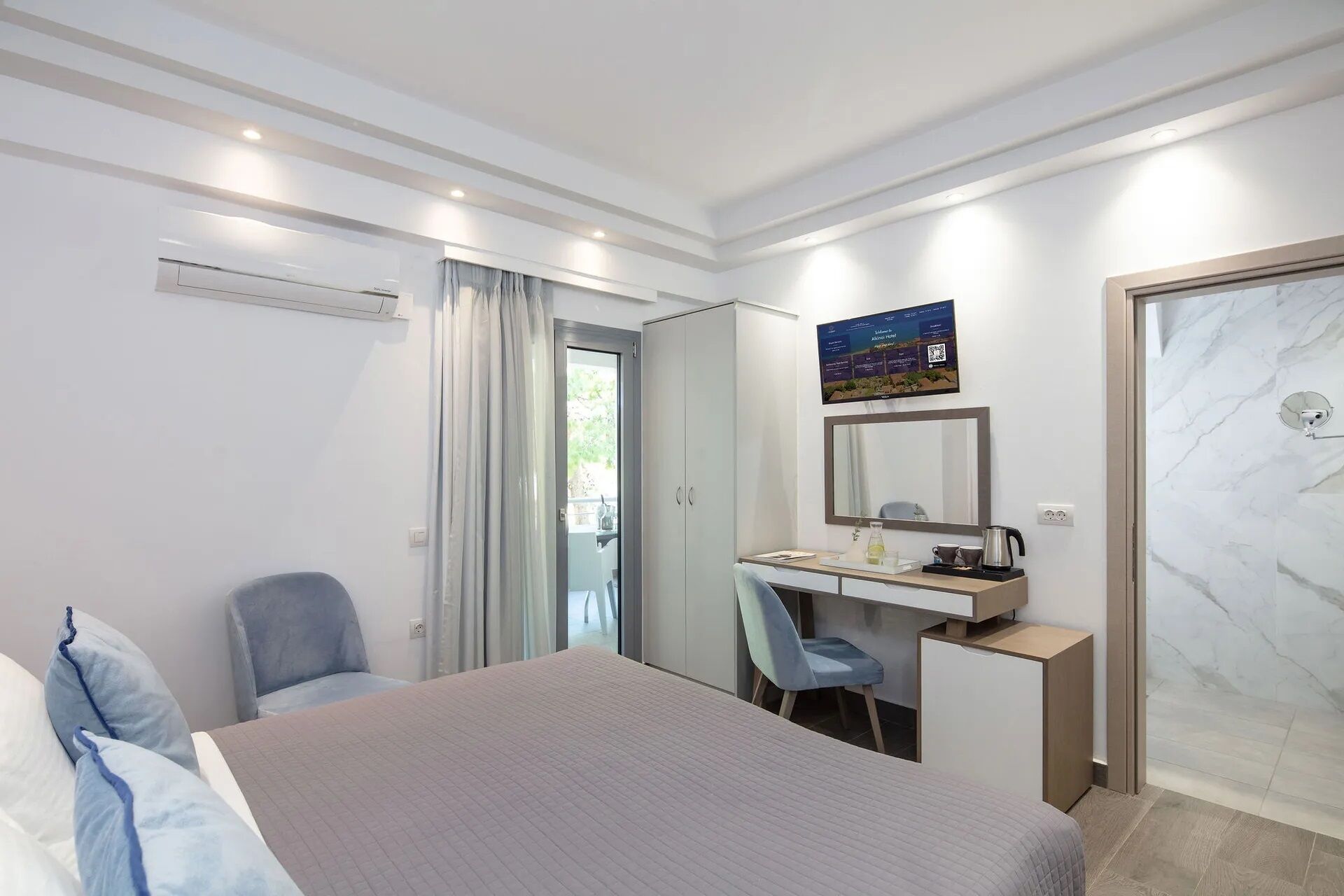 A modern hotel room with a bed, a desk, a chair, a wardrobe, a shower area, and a bathroom.